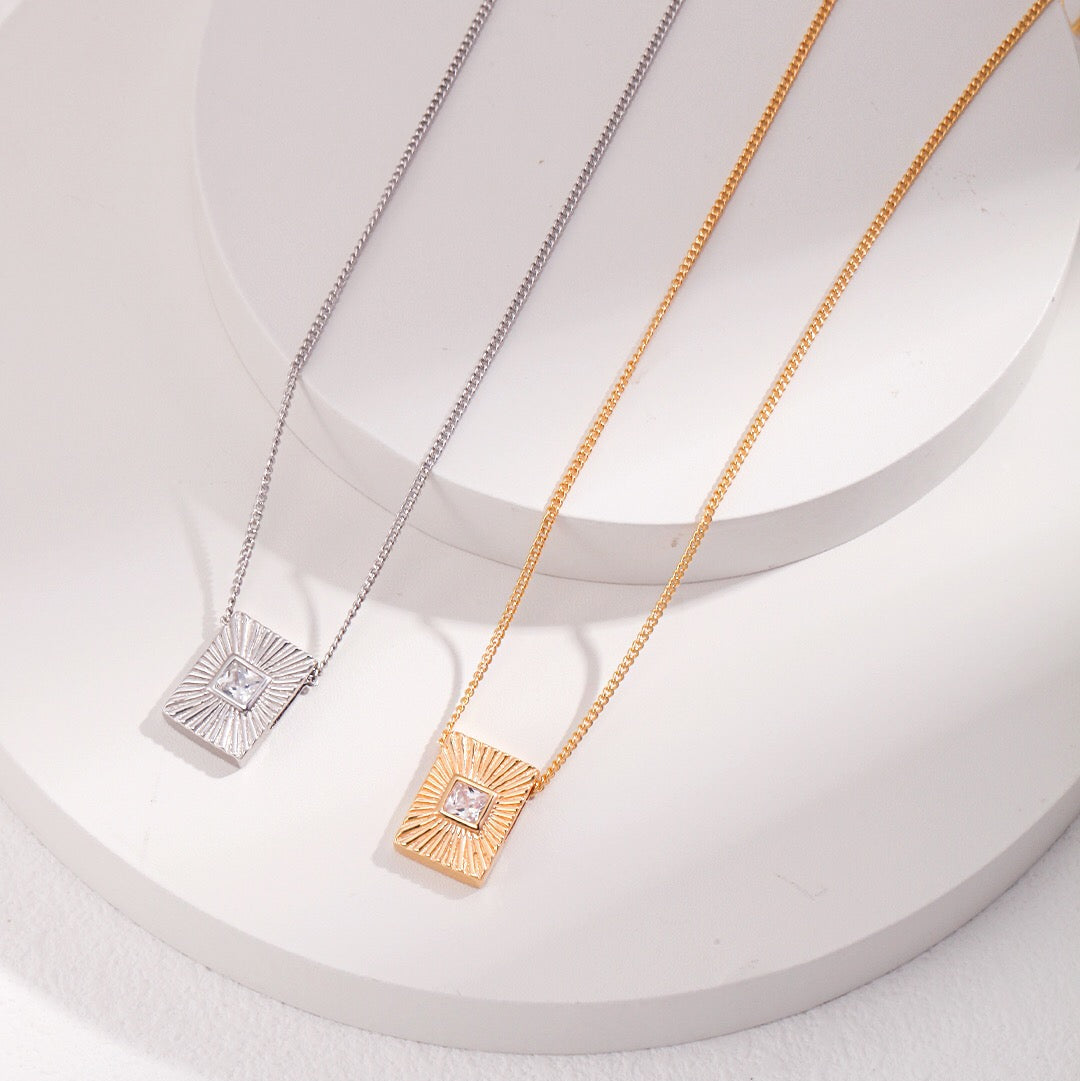 Two zircon necklaces, one with a silver chain and silver setting, and the other with a gold chain and gold setting, displayed on a white platform.