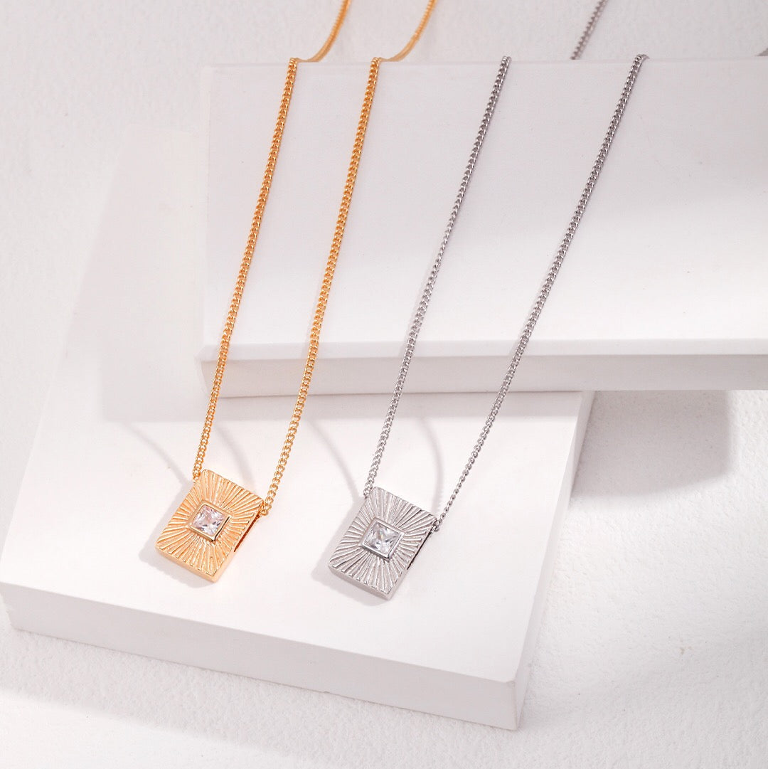 Two zircon necklaces, one with a silver chain and silver setting, and the other with a gold chain and gold setting, displayed on a white platform.